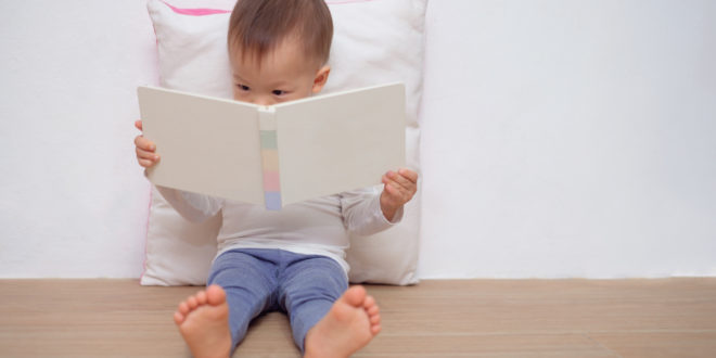 Cute little Asian 18 months / 1 year old toddler boy child sitting on floor, leaning against pillow, looking at a book