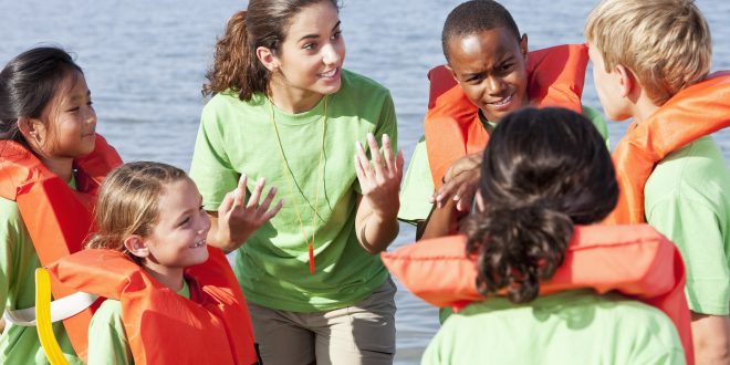 A camp counselor talking wearing a green shirt is talking to a group of children who are wearing life vests at camp.
