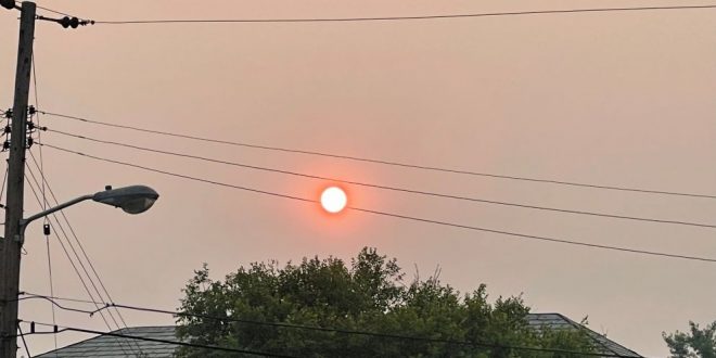 An orange-colored sun shines between the wires of a powerline in a hazy sky.