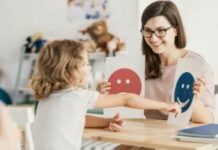 A child with autism looks at paper emoticons in a therapy session. The paper emoticons are held by a woman with dark hair. She is a therapist.