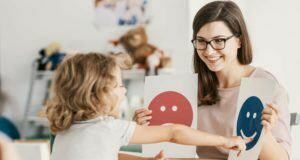 A child with autism looks at paper emoticons in a therapy session. The paper emoticons are held by a woman with dark hair. She is a therapist.