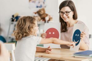 A child with autism looks at paper emoticons in a therapy session. The paper emoticons are held by a woman with dark hair. She is a therapist. 