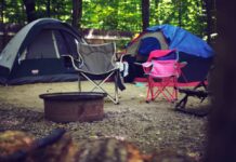 Stock photo of two portable camp chairs, one gray and one pink, in front of two tents on a campground. There is a firepit in front of the chairs.