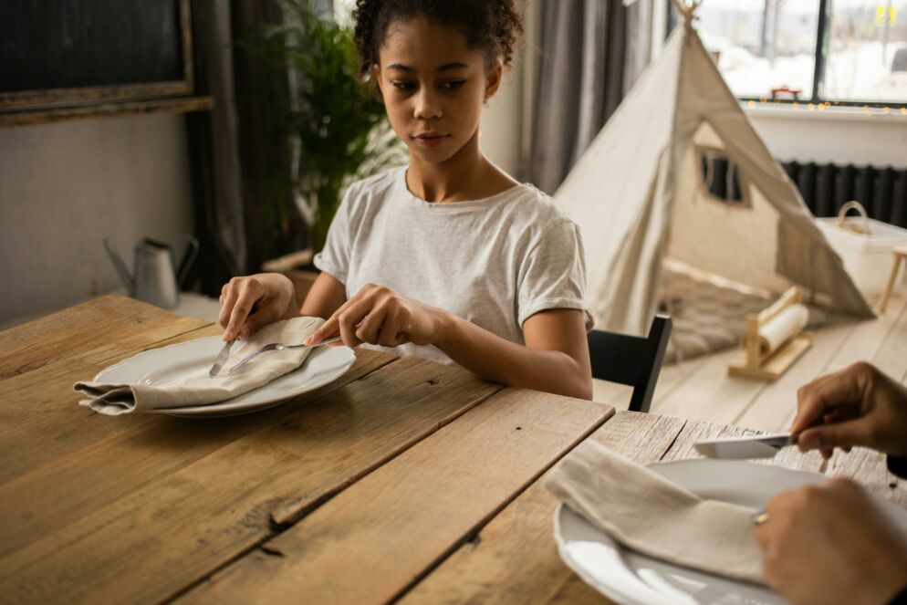 Young Black girl watches an adult use cutlery at a place setting as she tries to imitate.