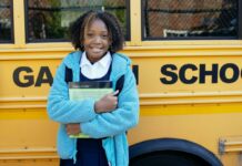 Get Ready for School with These 7 August Events Stock photo of a school-aged girl with dark curly hair standing in front of a yellow school bus. She is wearing a white-collared shirt under a turquoise jacket and she is holding a textbook to her chest and smiling at the camera.