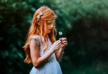 Stock photo of a young girl with long straight red hair holding a dandelion outside. She is wearing a pale blue dress with a matching bow headband.