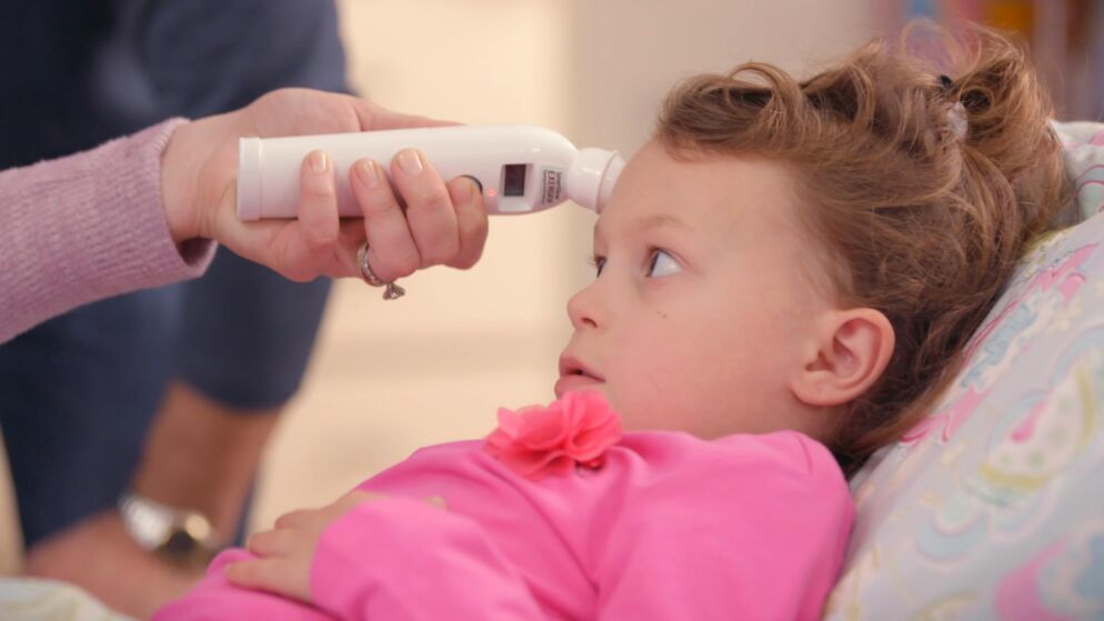 Little girl in a pink shirt lies down as a forehead thermometer is touched to her head to see if she has a temperature.