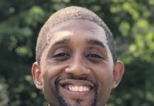 Photo of a man wearing a gray suit with a light blue tie. He is smiling at the camera, standing outside.