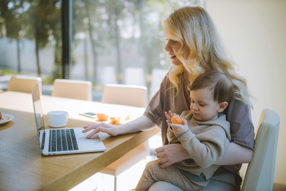 Woman holds a baby as she works on her laptop wiht sun streaming in from the window