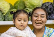 Mother smiles, holding child at farmers market with produce in the background behind them
