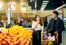 Poll: Where Do You Like to Grocery Shop As a Family? Two adults inspecting fruit at a grocery store with a young child in a cart next to them.