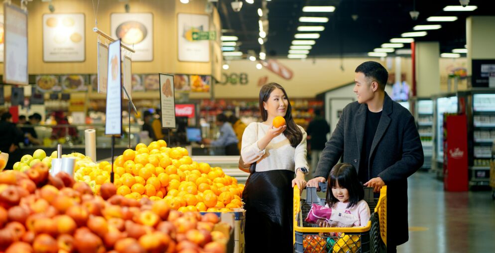 Two adults inspecting fruit at a grocery store with a young child in a cart next to them.