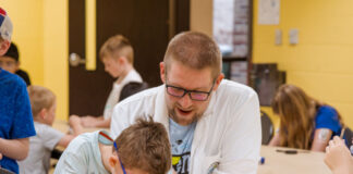 A scientist in a white lab coat supervises a young camper looking through a microscope.