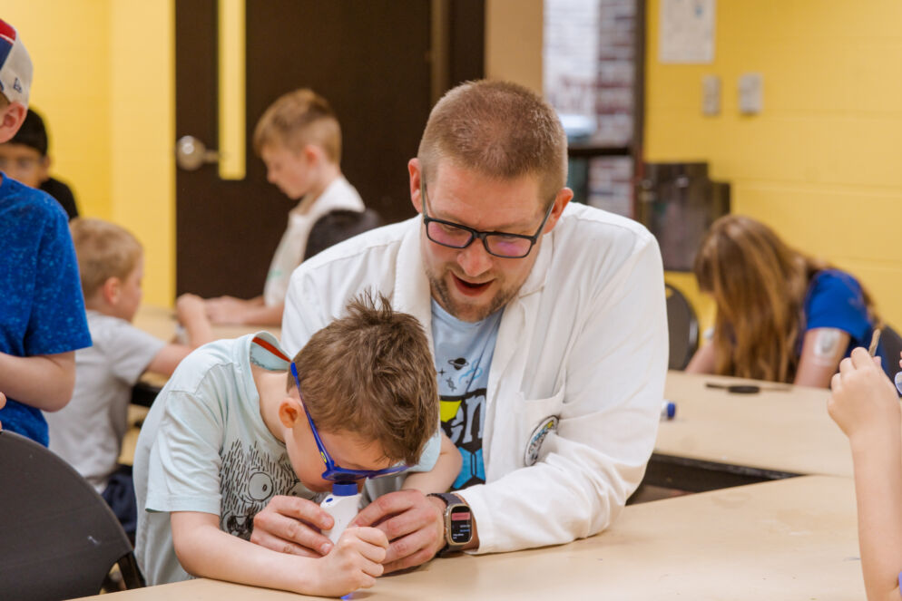 A scientist in a white lab coat supervises a young camper looking through a microscope.