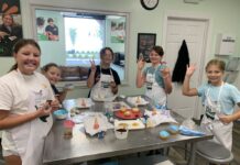 FREE Family Fun In Baltimore This September A group of kids having fun in the kitchen at Young Chefs Academy. They are standing around a table throwing up peace signs and waving at the camera.