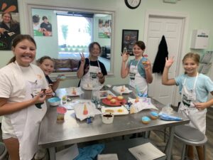 A group of kids having fun in the kitchen at Young Chefs Academy. They are standing around a table throwing up peace signs and waving at the camera.