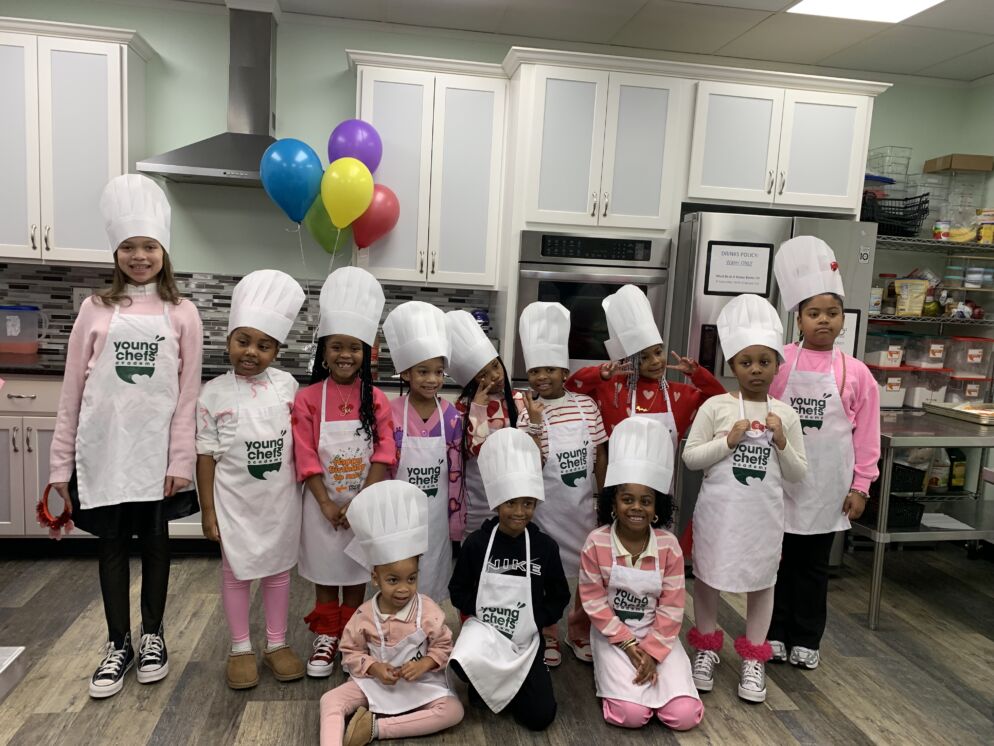 A group of kids from Young Chefs Academy pose for a photo wearing their white chef hats, a classic style, in the kitchen.