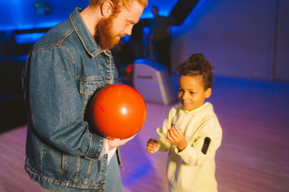 An adult man holds an orange bowling ball as a little girl watches