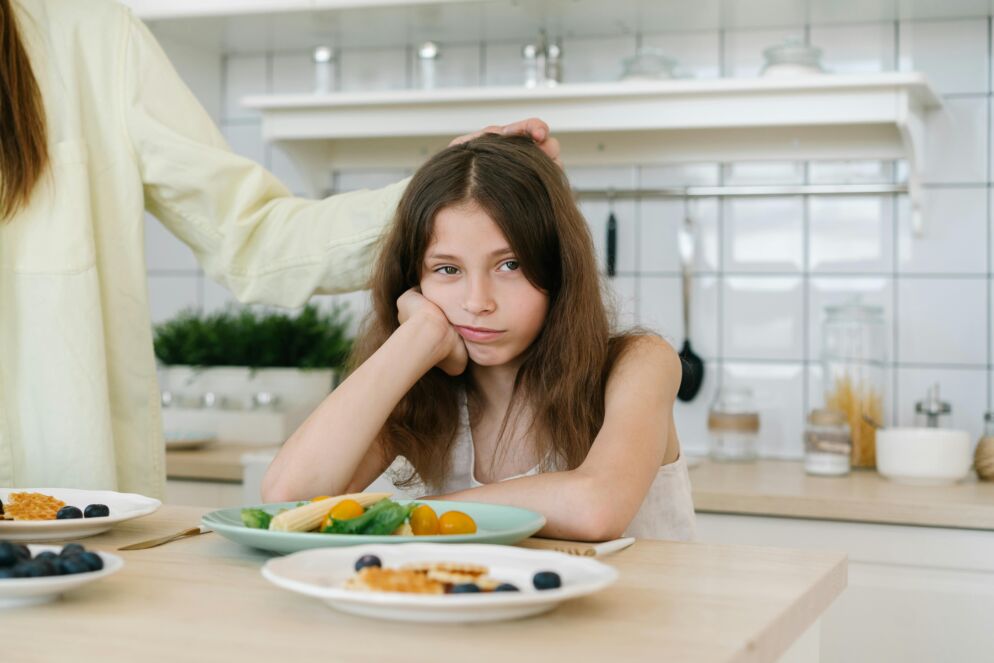 Girl sitting at a table with a plate of food, looking grumpy