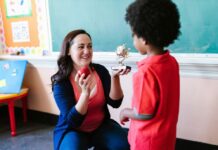 A teacher receives an apple and a trophy that says "best teacher" from a student