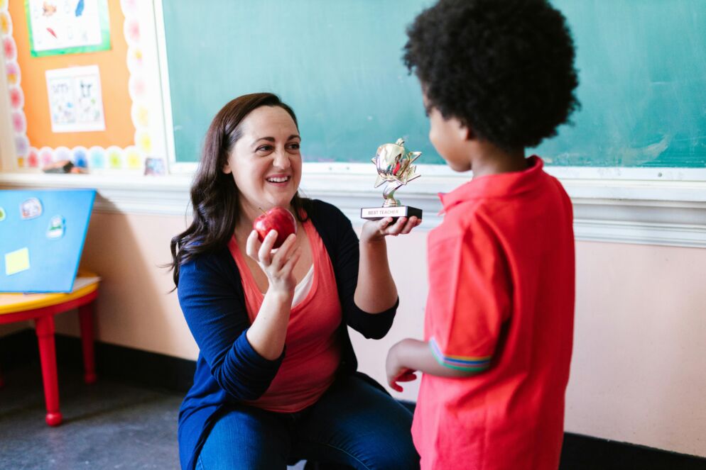 A teacher receives an apple and a trophy that says "best teacher" from a student