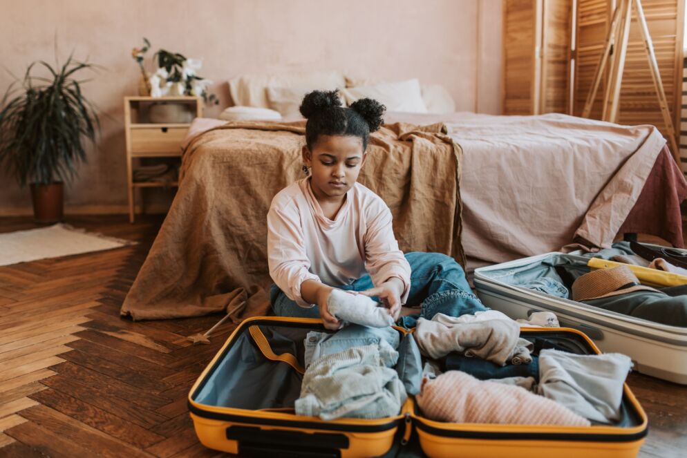 A child sits on the floor, looking pensive as she packs her suitcase. This photo represents packing for summer camp or a summer trip.