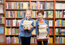 Make Reading Fun This Summer Kids standing in front of a bookshelf holding stacks of books
