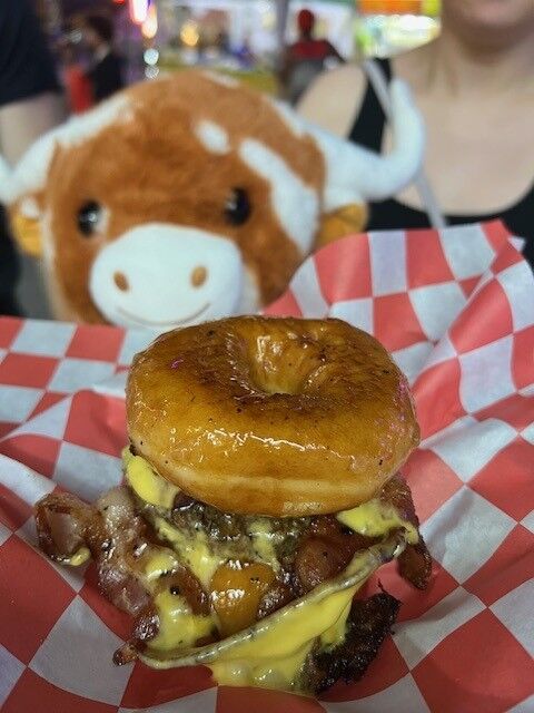 A stuffed cow looks at a burger sandwiched between two whole Krispy Kreme donuts, a sweet and savory treat at the Maryland State Fair