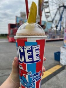 An Icee with a pickle spear stuck in the top held up against the backdrop of carnival rides at the Maryland State Fair