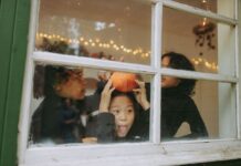 Three kids play by a window looking out on Halloween. One is holding a pumpkin and making a silly face.