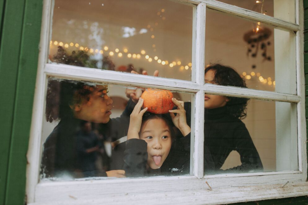 Three kids play by a window looking out on Halloween. One is holding a pumpkin and making a silly face.